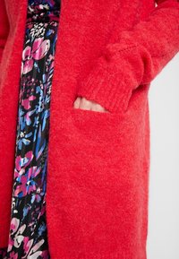 Röd stickad cardigan med en mjuk, texturerad yta, ribbade ärmslut och en framficka. Under syns en blommönstrad klänning.