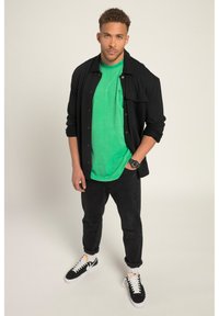 Camisa negra de manga larga, camiseta verde de manga corta, pantalones negros y zapatillas negras con suelas blancas, todo sobre un fondo neutral.