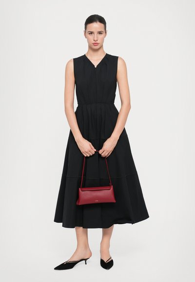 Vestido negro sin mangas con cintura ceñida y falda acampanada, combinado con un bolso rojo pequeño. Modelo lleva zapatos de punta negros.