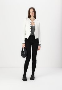 White cropped jacket with silver buttons, grey tank top, black skinny jeans, and black shoes. Holding a black, soft clutch handbag.