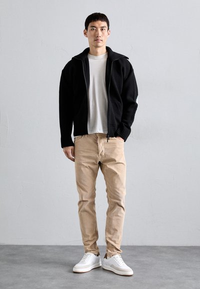 Homme standing contre un mur plain, portant une veste zippée noire, un t-shirt blanc, un pantalon beige et des baskets blanches, les mains dans les poches.