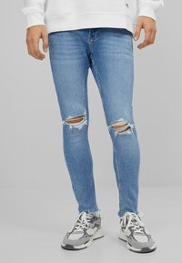 Jeans ajustados de mezclilla azul claro con desgastes en los dobladillos y dos desgastes en las rodillas, combinados con zapatillas grises y blancas sobre un fondo neutro.