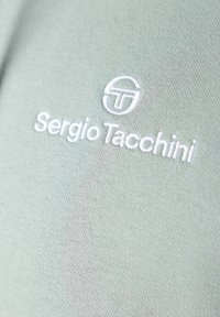 Tessuto verde con una trama liscia presenta la scritta bianca ricamata "Sergio Tacchini" e un logo circolare.
