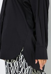 Schwarzes Hemd mit gebogenem Saum, langen Ärmeln mit Knopfdetail und glatter Stoffstruktur, kombiniert mit gemusterten, hoch taillierten Hosen.