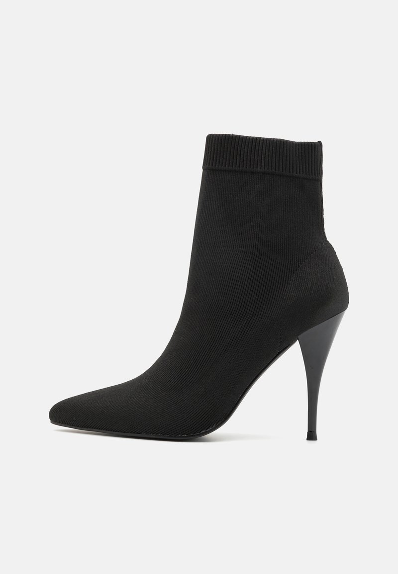 Bottine chaussette noire en maille avec bout pointu et talon haut fin sur fond blanc.