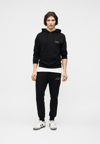 Νέος άντρας όρθιος, φοράει μαύρο φούτερ με κουκούλα, μαύρο παντελόνι φόρμας με λευκή επιγραφή, λευκό μπλουζάκι εσωτερικά και λευκά αθλητικά παπούτσια με μαύρες ρίγες.