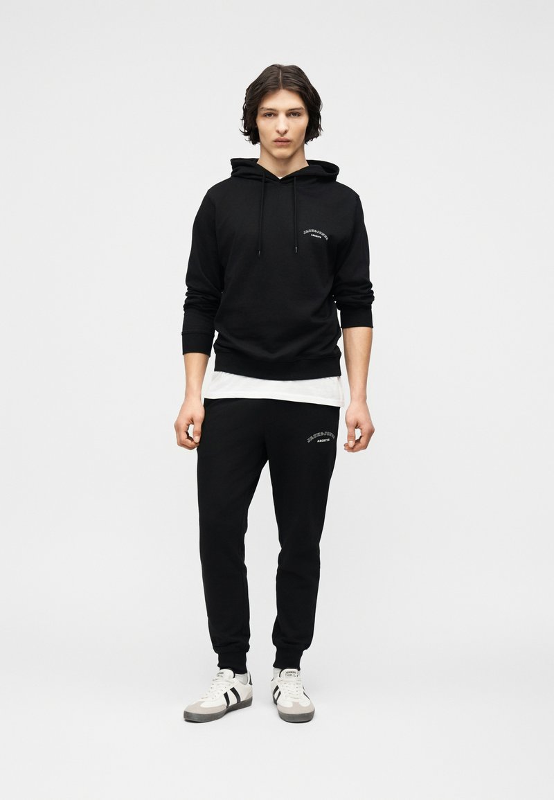 Νέος άντρας όρθιος, φοράει μαύρο φούτερ με κουκούλα, μαύρο παντελόνι φόρμας με λευκή επιγραφή, λευκό μπλουζάκι εσωτερικά και λευκά αθλητικά παπούτσια με μαύρες ρίγες.