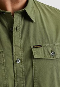 Camicia verde oliva con bottoni, colletto, tasche sul petto e un'etichetta nera con testo giallo che recita "PME Legend". La texture sembra liscia.