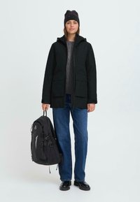 Chaqueta parka negra con cremallera en el frente, grandes bolsillos y capucha, combinada con un suéter gris y jeans azules. Mochila negra con correas ajustables.