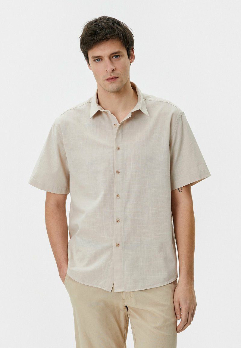 Camisa beige de manga corta con cierre de botones en el frente, tela ligeramente texturizada y cuello clásico; cuenta con botones opacos y un ajuste relajado.