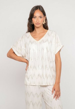 Femme aux longs cheveux noirs portant un haut à manches courtes à motif zigzag blanc et or et un pantalon assorti, debout avec une main sur la hanche.