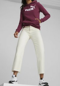 Bordeaux hættetrøje med hvid "PUMA" logo, cremefarvede cropped jeans med frynset kant, og sort-hvide sneakers. Glat tekstur og afslappet design.