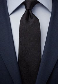 Svart paisleymönstrad slips i texturerad tyg, med ett subtilt mönster, bärs med en mörkblå kostym och en ljusblå skjorta.