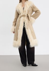 Cappotto in ecopelliccia beige con colletto e polsini in pelliccia morbida, vita cintata e bordo in crema a contrasto. Abbinato a pantaloni neri a gamba larga e scarpe a punta.