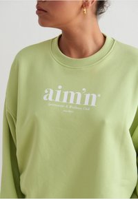 Svetlozelená mikina vyrobená z mäkkej tkaniny, s rebrovaným výstrihom a bielym textom "aim'n Sportswear & Wellness Club since 2013."