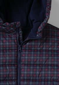 Giacca imbottita con motivo trapuntato in blu navy, rosso e grigio. Presenta una chiusura con zip e un rivestimento morbido per il cappuccio. Tessuto strutturato per il calore.
