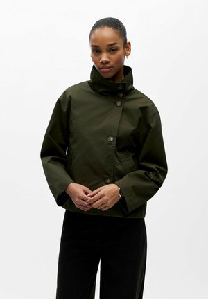 Femme portant une veste boutonnée à col montant vert foncé et un pantalon noir, debout les mains jointes sur un fond blanc.