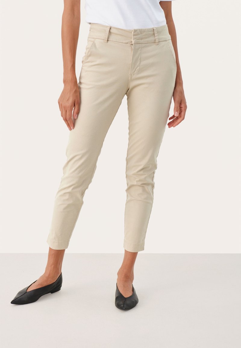 Pantalons beiges ajustés avec une texture lisse, des jambes fuselées et une taille mi-haute, associés à des chaussures plates noires.