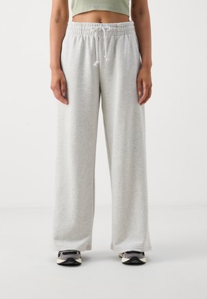 Persoon die lichtgrijze sweatpants met wijde pijpen draagt, voorzien van een elastische tailleband en trekkoord, gecombineerd met grijze en zwarte sneakers.