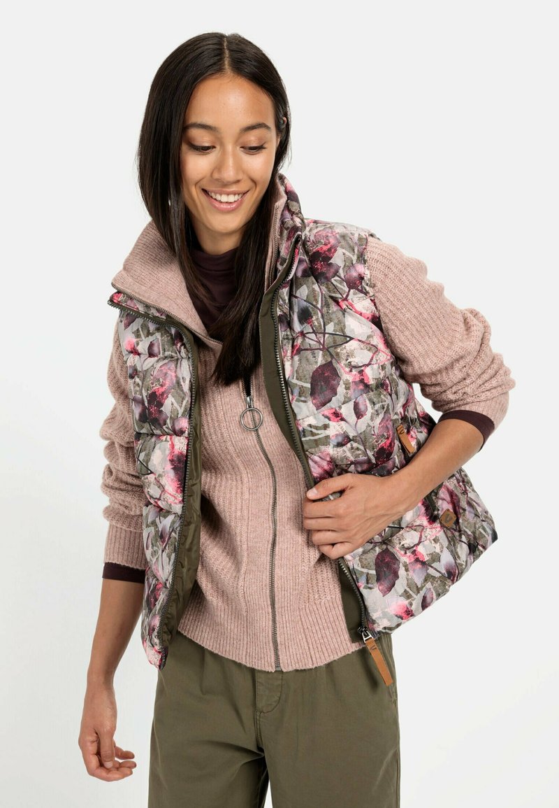 Blomsterpræget vest med quiltet tekstur i grønne og lyserøde farver, lagdelt over en lys pink, ribbet langærmet top. Jordfarvede bukser.