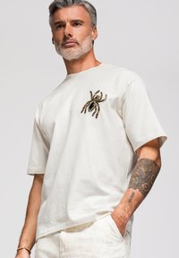 Homme aux cheveux gris et à la barbe porte un t-shirt blanc avec une grande araignée décorative sur la poitrine et a l’avant-bras droit tatoué.