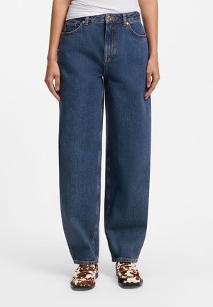 Persona che indossa jeans blu scuro larghi e scarpe bianche e marroni con motivo, in piedi su sfondo bianco.