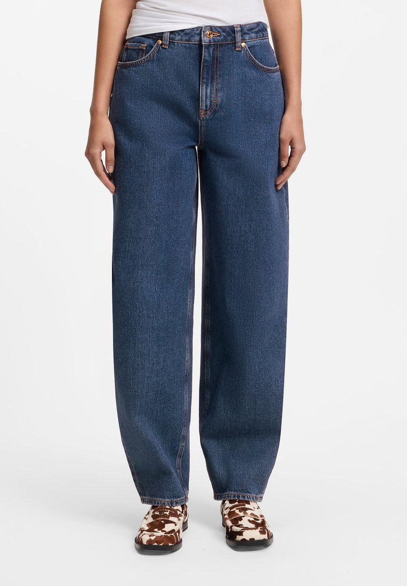 Personne portant un jean bleu foncé ample et des chaussures blanches et marron à motifs, debout sur un fond blanc.