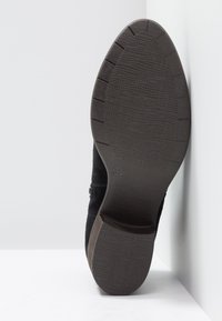 Musta mokkakenkä, jossa on teksturoitu kumipohja, harvennettu muotoilu ja hienovaraiset ompeluyksityiskohdat. Koon merkintä näkyvissä pohjassa.