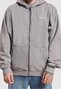 Persona vestida con una sudadera gris con cremallera y bolsillos frontales, jeans azules, tatuajes visibles en las manos y anillos en los dedos.