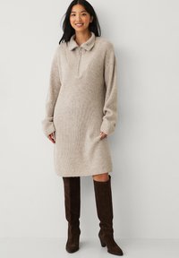 Beige gebreide jurk met een kraag en halve rits, met lange mouwen en een relaxte pasvorm. Gecombineerd met hoge bruine suède laarzen.