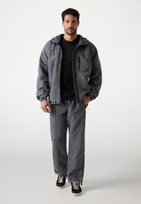 Chaqueta gris con capucha y cremallera, combinada con pantalones grises a juego. Camiseta negra por debajo, zapatillas negras y beige. Corte relajado, tejido texturizado.