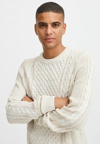 Suéter de punto color crema con patrones de trenzado texturizados, un cuello redondo acanalado, y puños anchos, con un corte holgado.