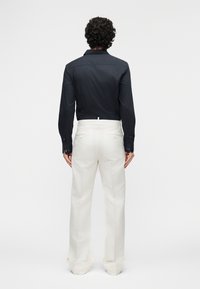 Homme aux cheveux bouclés portant une chemise noire ajustée à manches longues et un pantalon blanc ample taille haute, debout de dos.