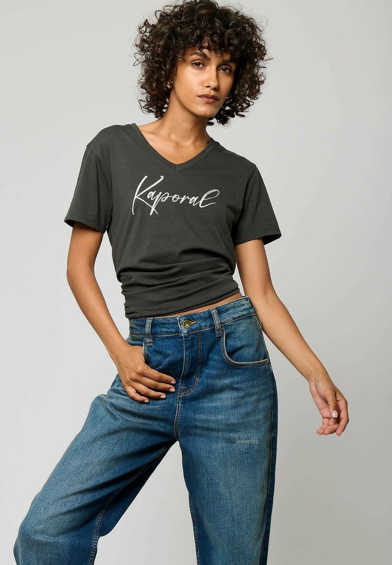 T-shirt gris foncé à col en V avec le texte "Kaporal" blanc, associé à un jean en denim bleu à coupe loose avec des boutons et des poches avant.