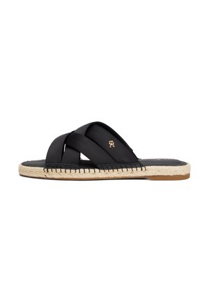 Sandale slide noire avec des brides croisées, semelle en jute tissée et petit logo doré sur la bride, vue de côté sur fond blanc.