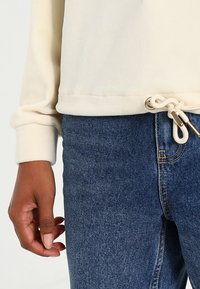 Lys beige hettegenser med snøring i kanten, kombinert med blå denim jeans. Synlig tekstur i stoffet og gyldne metalldetaljer.