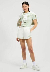 Mujer de pie con una camisa de manga corta verde claro, pantalones cortos blancos, zapatillas blancas y una sudadera blanca-verde drapeada sobre los hombros.