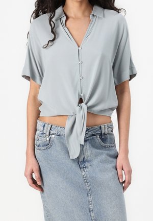 Femme portant une chemise à manches courtes bleu clair nouée à la taille, associée à une jupe en jean bleu clair.