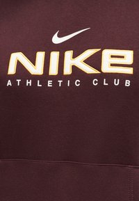 Σκούρο μπορντό ύφασμα με μεγάλο λευκό κείμενο "NIKE" με χρυσό περίγραμμα, λευκή σφήνα Nike πάνω από αυτό και μικρότερο λευκό κείμενο "ATHLETIC CLUB" από κάτω.