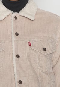 Beige kordjacka med en fluffig vit krage, med bruna knappar och en röd Levi's-tag på vänster ficka. Texturerad med vertikala ribbor.