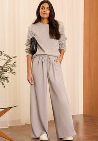 Sivý pletený sveter s voľným strihom, svetlosivé široké nohavice s viazačkou v páse, spárované s bielymi teniskami a čiernou kabelkou.
