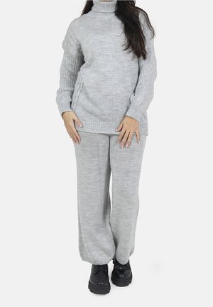 Femme portant un pull à col roulé tricoté gris clair et un pantalon large assorti avec des chaussures à plateforme noires, debout devant un fond blanc.