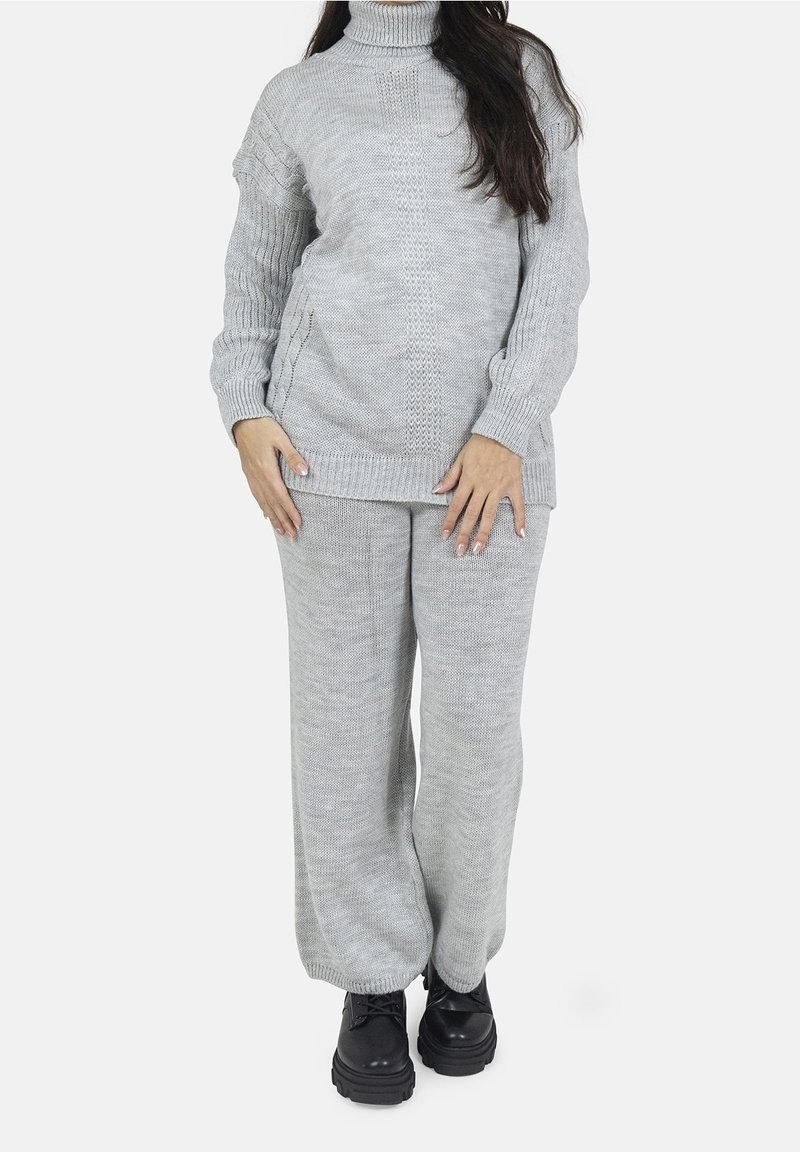 Femme portant un pull à col roulé tricoté gris clair et un pantalon large assorti avec des chaussures à plateforme noires, debout devant un fond blanc.