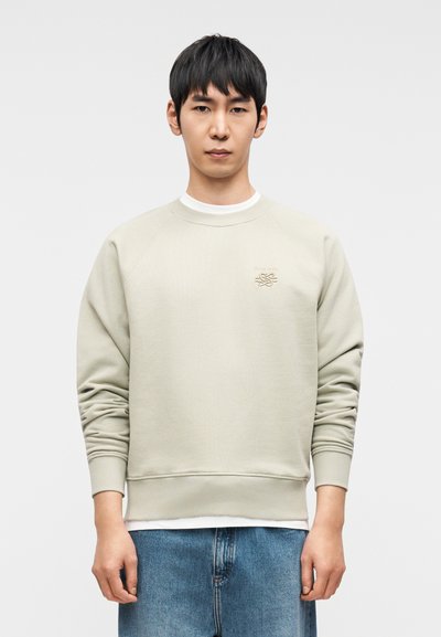 Ανοιχτό γκρι φούτερ με στρογγυλή λαιμόκοψη και ριμπ μανσέτες, με μικρό κεντημένο λογότυπο στο στήθος. Φορεμένο πάνω από ένα λευκό πουκάμισο.