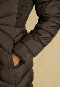 Čierna páperová bunda s diagonálnym prešívaním, ktorá má zipsovú kapsu a na rukáve je embosovaný názov značky 'Joules'.