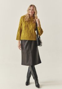 Senfgelber Strickpullover mit hohem Kragen, kombiniert mit einem dunklen Leder-Midirock und kniehohen schwarzen Stiefeln. Schwarze Umhängetasche.