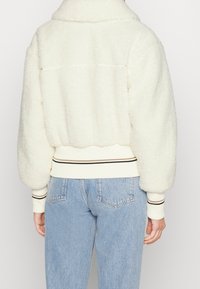 Person med krämfärgad fluffig jacka med randiga muddar och nederkant, matchat med ljusblå denimjeans, sedd bakifrån.