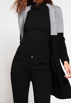 Vrouw met een zwarte coltrui, zwarte broek en een lange grijs-zwarte colorblock cardigan tegen een egale achtergrond.