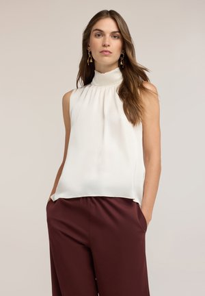 Vrouw die een mouwloze witte blouse met hoge hals en donkerrode broek draagt, met handen in de zakken tegen een effen achtergrond.