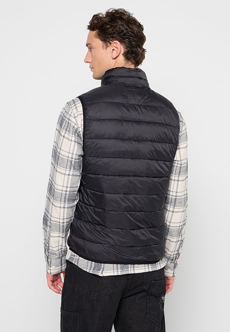 Svart pufferväst med horisontell vaddering, hög krage och en slät, blank yta, bärs över en grå och vit rutig skjorta.